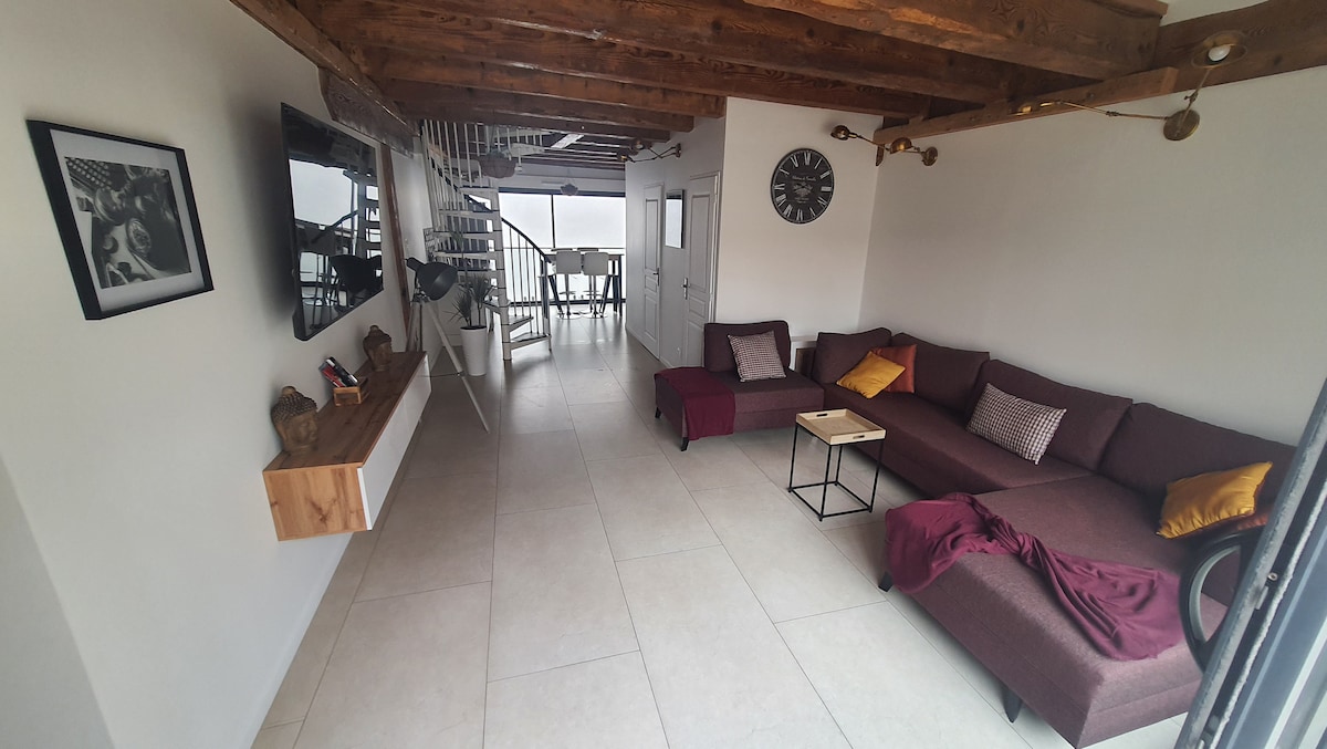 A spacious living area features a deep pink sectional sofa accented with cushions. A light wooden coffee table is positioned nearby, while a flat-screen TV is mounted on the wall. Large windows allow natural light to fill the space, contributing to an airy atmosphere.
