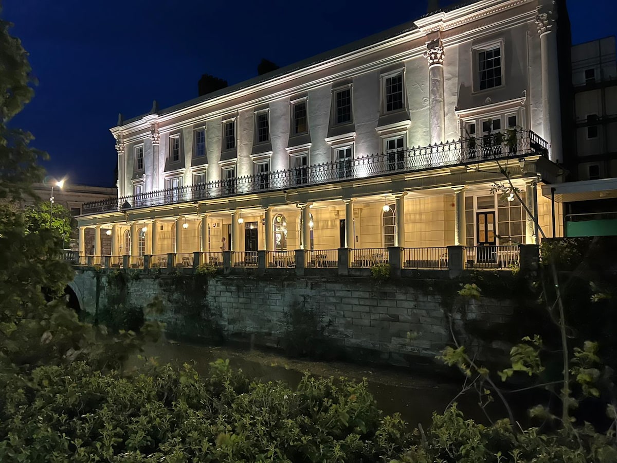 The historic Grade II Listed Victoria Colonnade is illuminated at night, showcasing its elegant facade and decorative architectural details. The structure features balconies and large windows, while reflections shimmer on the river below, creating a tranquil nighttime scene.