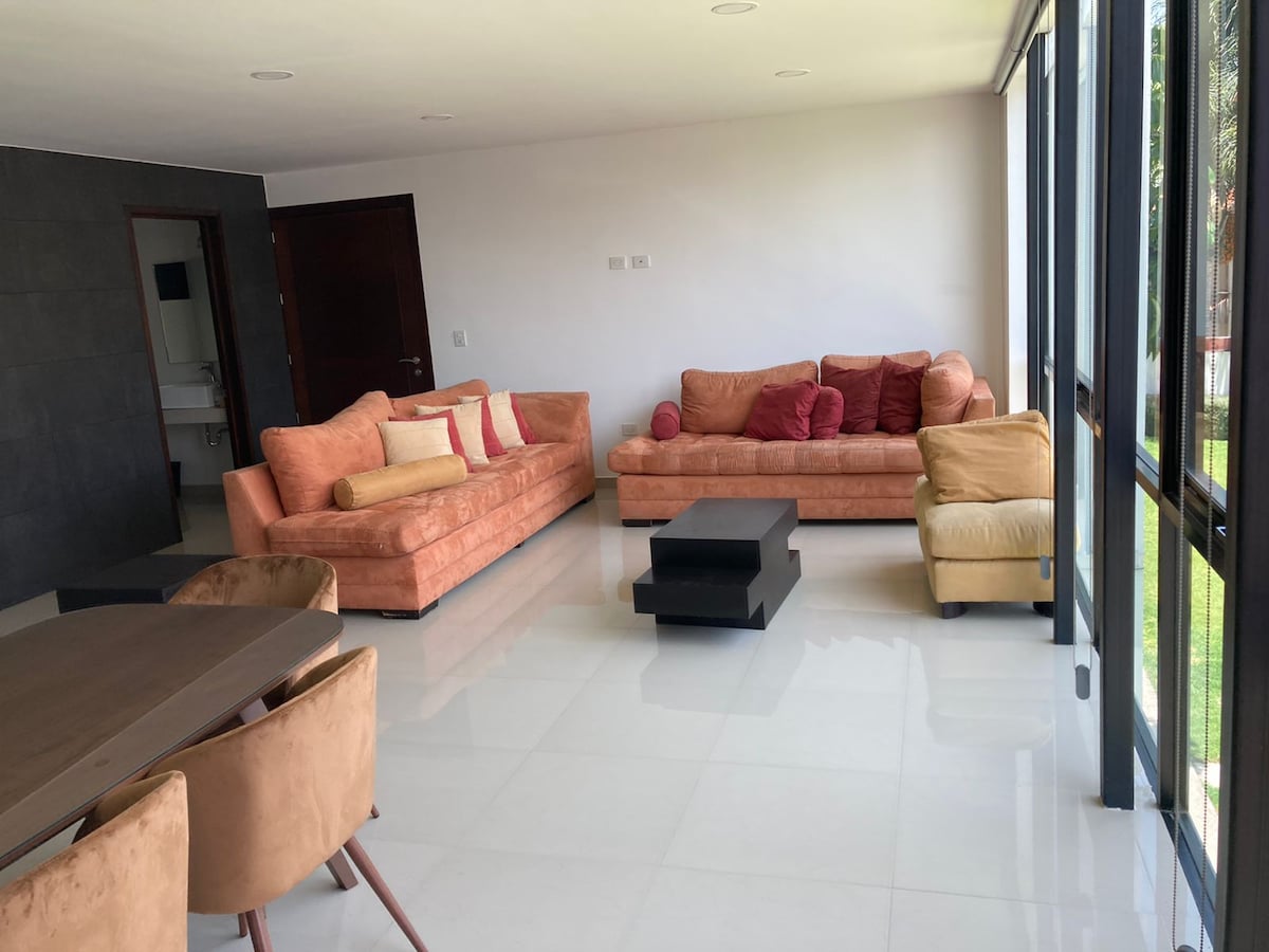 A spacious living area features large glass windows allowing natural light. Two comfortable sofas are arranged with accent pillows, complemented by a low coffee table. A dining table with chairs is positioned nearby, emphasizing the open layout and connection to the outdoors.