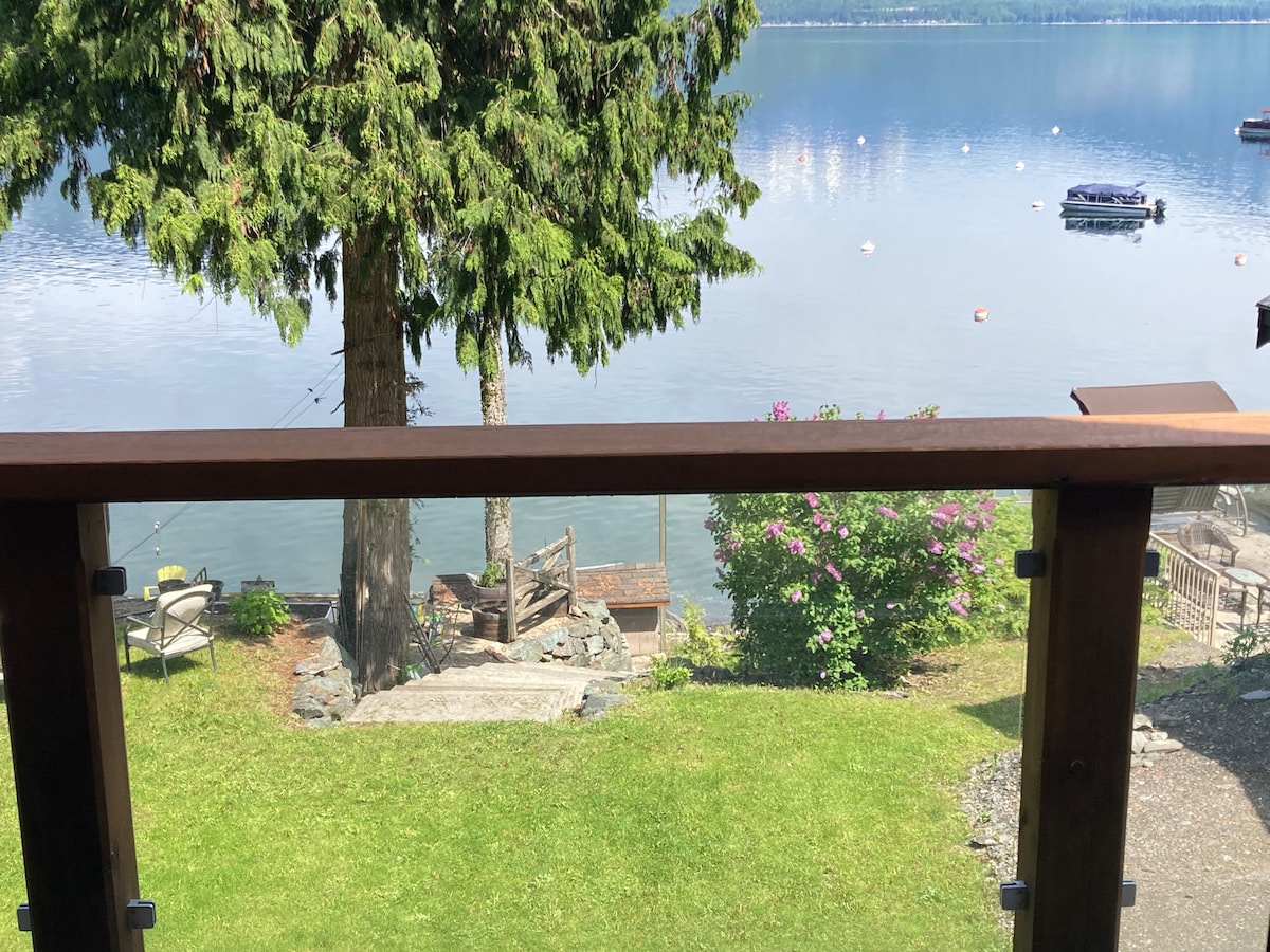 A private balcony overlooks a serene lake setting, framed by lush greenery and a tall tree. Soft grass leads down to the water, where boats are anchored, and colorful flowers add a touch of vibrancy to the landscape.