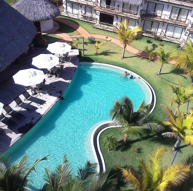 A curvy swimming pool is surrounded by lush green grass and tropical palm trees. Several sun loungers and umbrellas are positioned on a wooden deck, offering shaded relaxation areas. Two individuals can be seen enjoying the water, enhancing the inviting ambiance.
