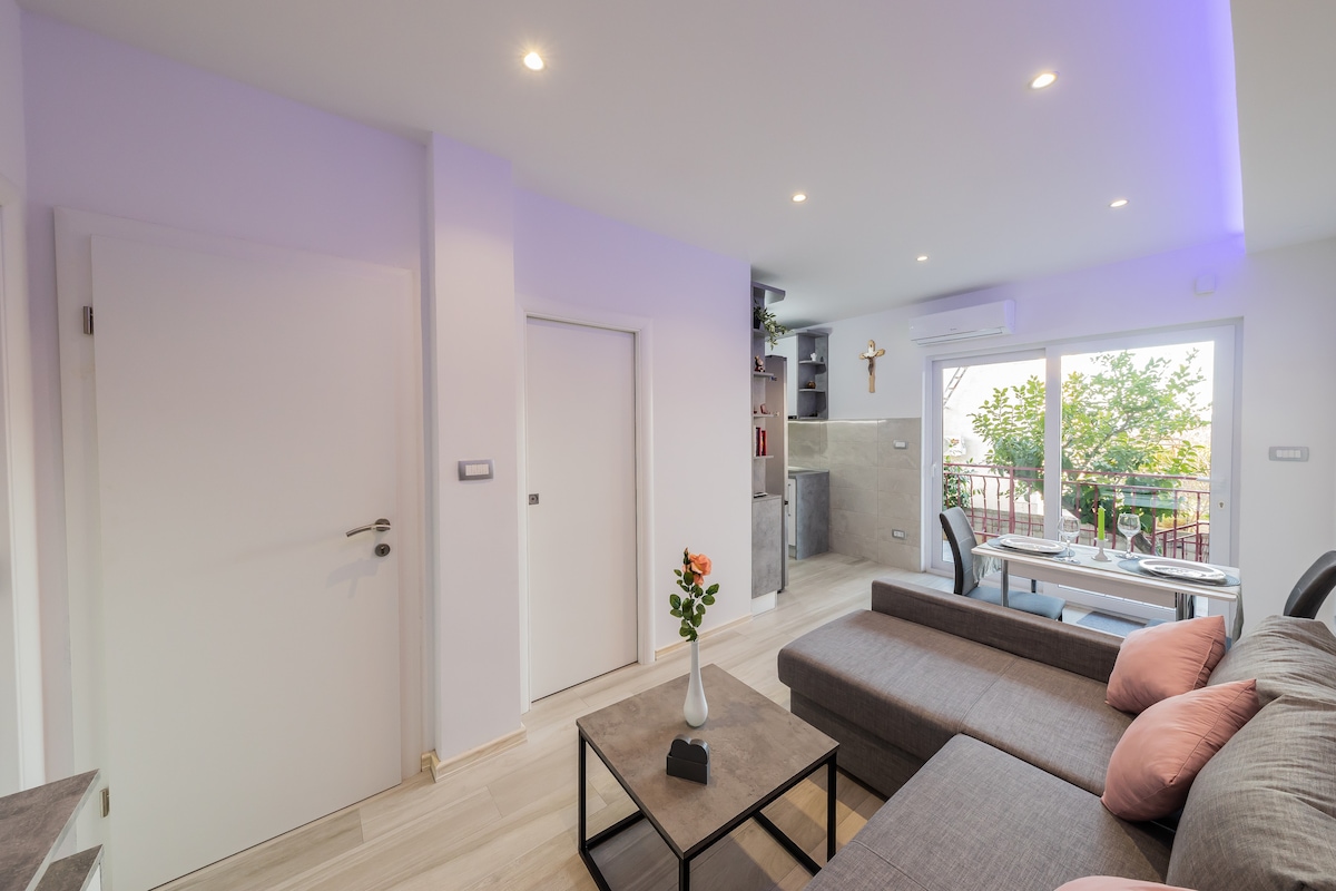 A modern living space showcases a grey sectional sofa adorned with soft pink cushions. Natural light fills the room through large glass doors that open to a balcony, while a small dining table and a decorative vase with flowers add visual interest.