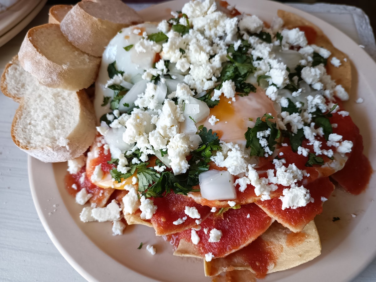 A plate of chilaquiles is presented with crispy tortilla chips layered beneath a rich red sauce. A sunny-side-up egg rests on top, accompanied by diced onions, fresh cilantro, and crumbled cheese. Slices of bread are arranged neatly beside the dish.