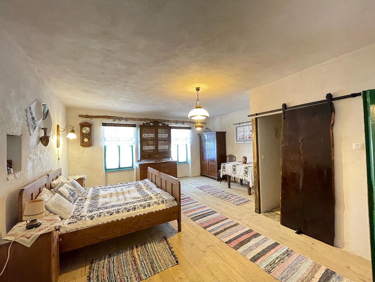 A spacious guest room features a wooden bed with decorative pillows, surrounded by a rustic wooden wardrobe and a smaller table set with two chairs. Natural light enters through two windows, while a patterned rug adds warmth to the wooden floor.