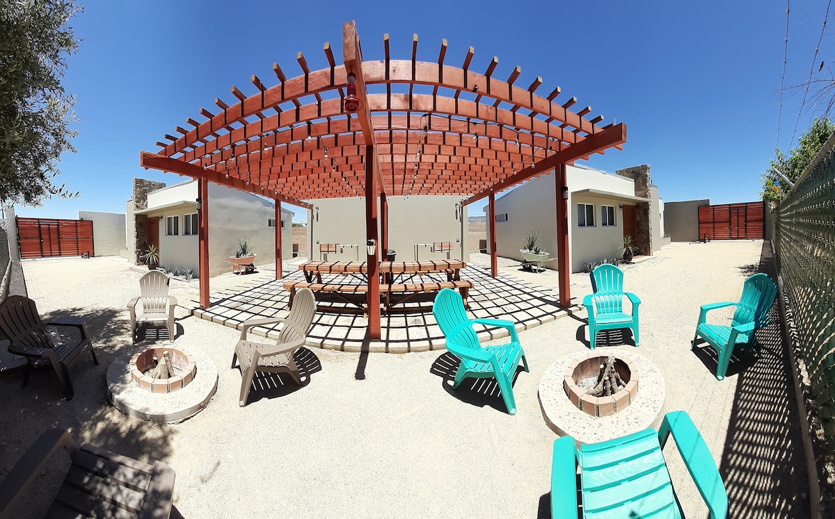 An outdoor space is shown with a wooden pergola providing shade over a large circular table surrounded by several brightly colored chairs. A firepit is positioned on the ground, and the surrounding area features gravel landscaping and two buildings in the background.