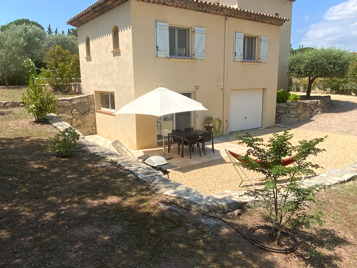 An outdoor area is featured, showcasing a stone pathway leading to a patio with a table and chairs under a large umbrella. Two lounge chairs are positioned nearby, and greenery surrounds the space, enhancing a tranquil and natural environment.