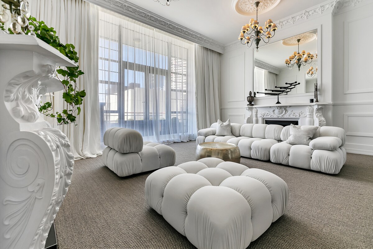 The living area features a soft, white seating arrangement that promotes relaxation. Large windows allow ample natural light to fill the room, highlighting the elegant chandelier and decorative elements. Subtle textures enrich the carpeted flooring, contributing to a sophisticated ambiance.