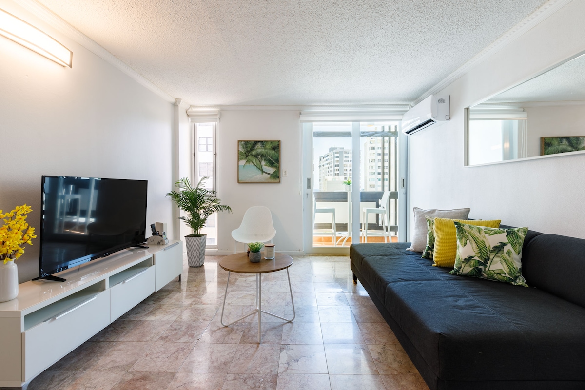 A spacious family room features a large sectional sofa with decorative pillows and a coffee table. A 50-inch TV is positioned on a media console. Natural light enters through sliding glass doors leading to a balcony, with greenery adding a comforting touch to the space.