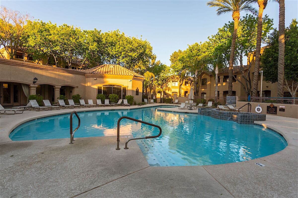 A resort-style pool is featured, framed by lush palm trees and shaded lounge chairs arranged around the perimeter. The pool has a gentle curve, with steps leading into the clear water. Nearby, a hot tub is visible, surrounded by stone accents.