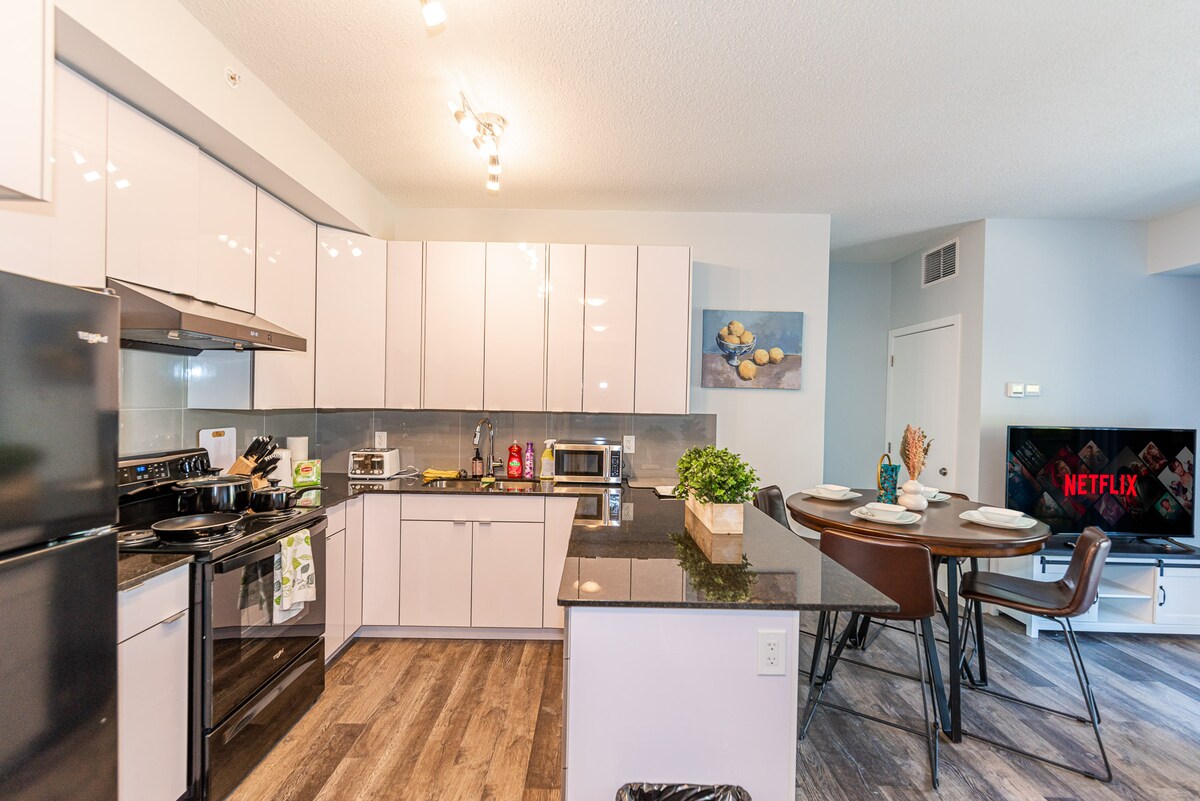 A modern kitchen features sleek, high-gloss cabinetry and granite countertops. Stainless steel appliances, including an oven, are visible, along with dishware displayed on the counter. A dining table is set for four, with a smart TV positioned nearby, offering entertainment options.