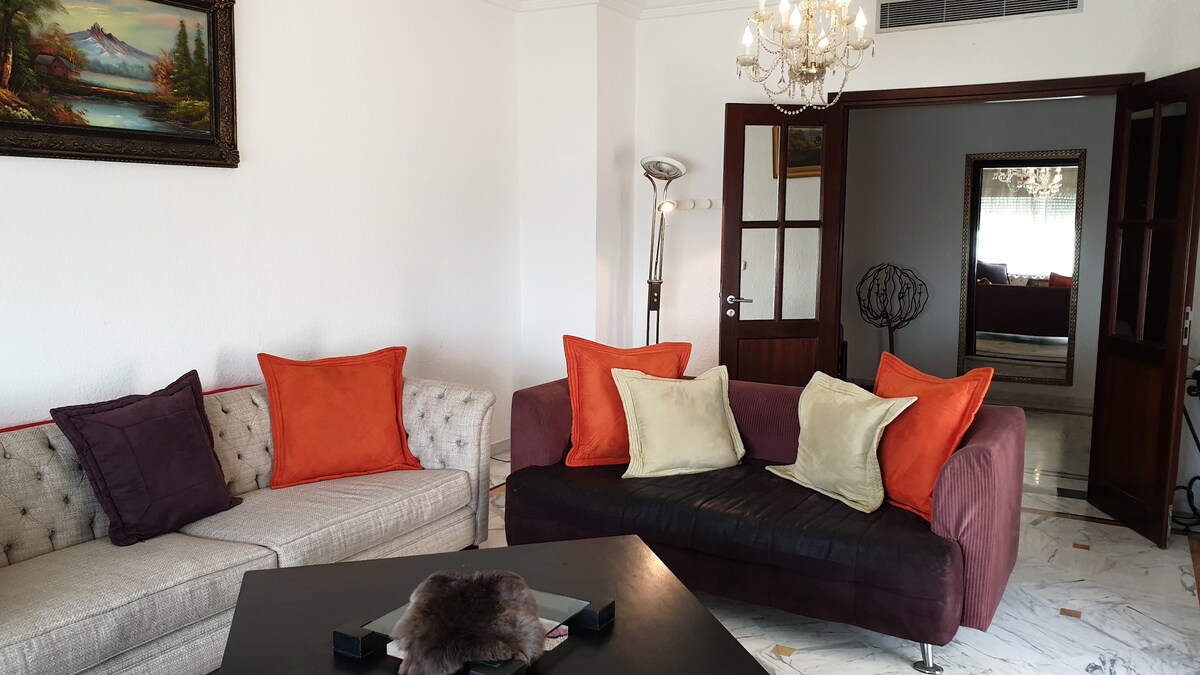 A living area features a mix of seating options, including a light-toned sofa with button detailing and a dark, plush couch adorned with colorful throw pillows. A coffee table is centered on the marble floor, and a chandelier adds soft illumination. A mirror and additional room can be seen in the background.