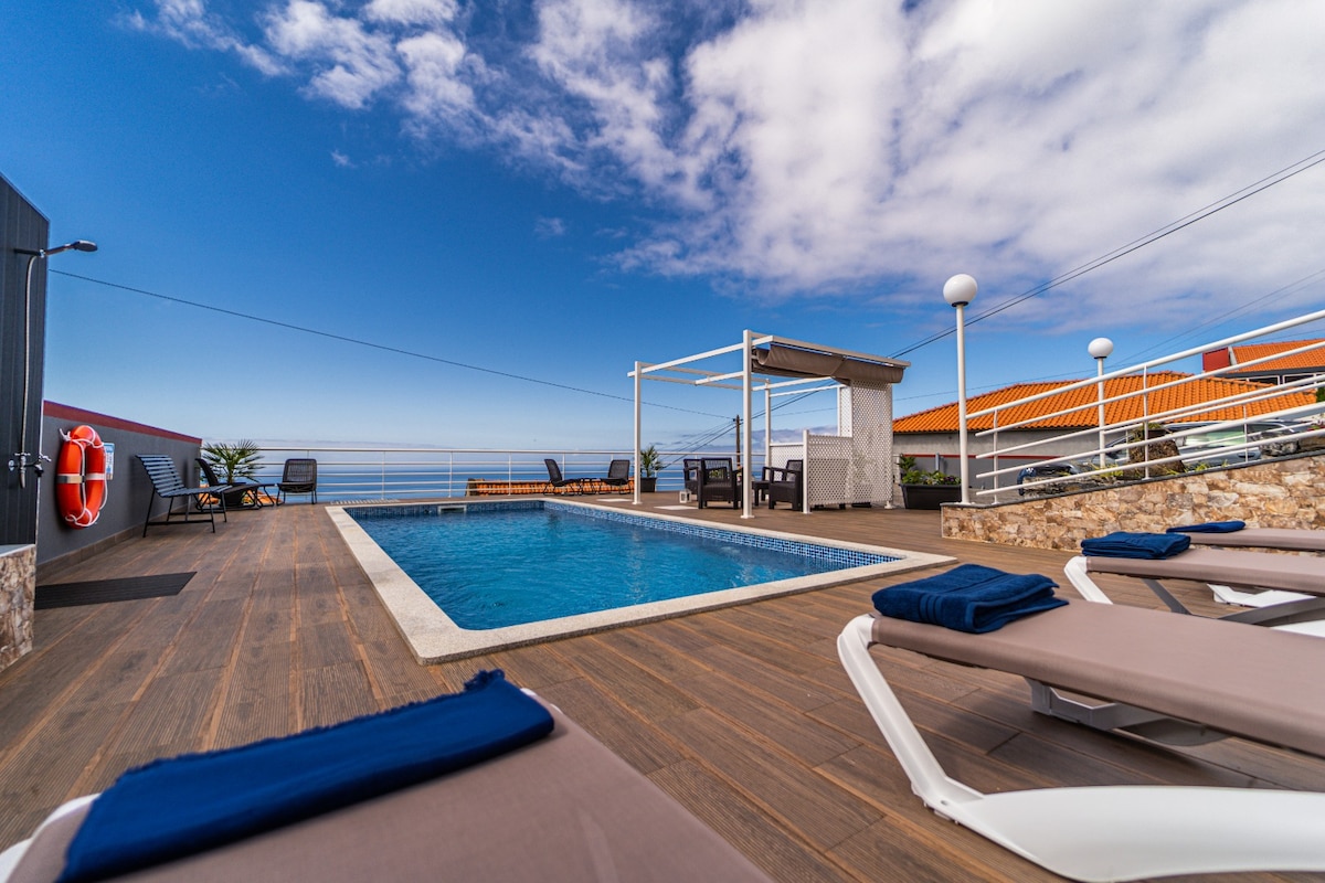 A swimming pool is framed by wooden decking, offering lounge chairs with navy-blue towels. A shaded seating area is visible in the background, accompanied by scenic views of the horizon and blue sky. The atmosphere suggests a peaceful outdoor space for relaxation.