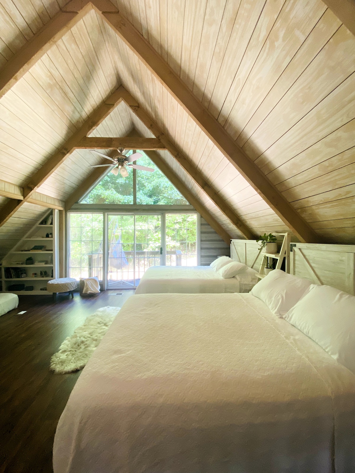 A spacious loft bedroom features two queen beds with soft bedding and an open, airy design. Large windows provide natural light and views of the surrounding trees, enhancing the serene atmosphere. A comfortable seating area is visible, creating a welcoming retreat.