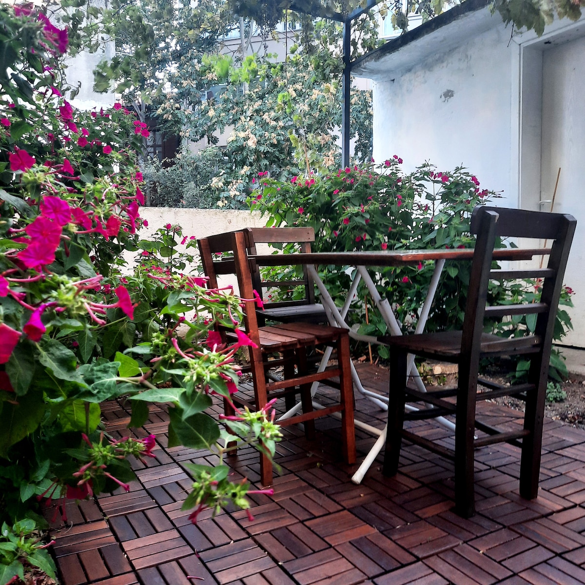 A small outdoor garden area features a wooden patio with three chairs and a table positioned among vibrant flowering plants. Lush greenery surrounds the space, creating a private and serene environment for relaxation.