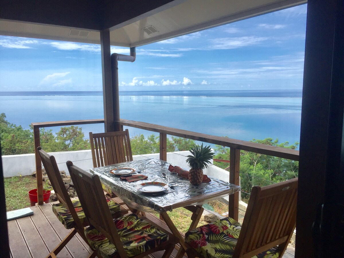 A dining area features a wooden table set for four, with colorful seat cushions. The spacious balcony offers views of the tranquil lagoon and sky, while tropical greenery frames the scene. Natural light fills the space, highlighting the inviting outdoor dining experience.