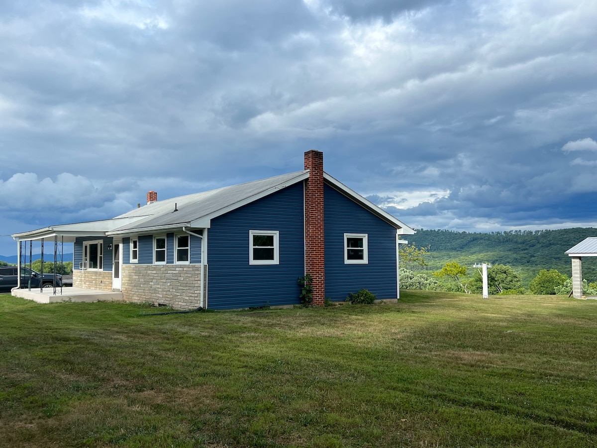 A spacious blue house with a stone foundation is set against a backdrop of rolling hills. The structure features multiple windows and a chimney, while a covered patio extends from one side, offering outdoor seating options. Green grass surrounds the home.
