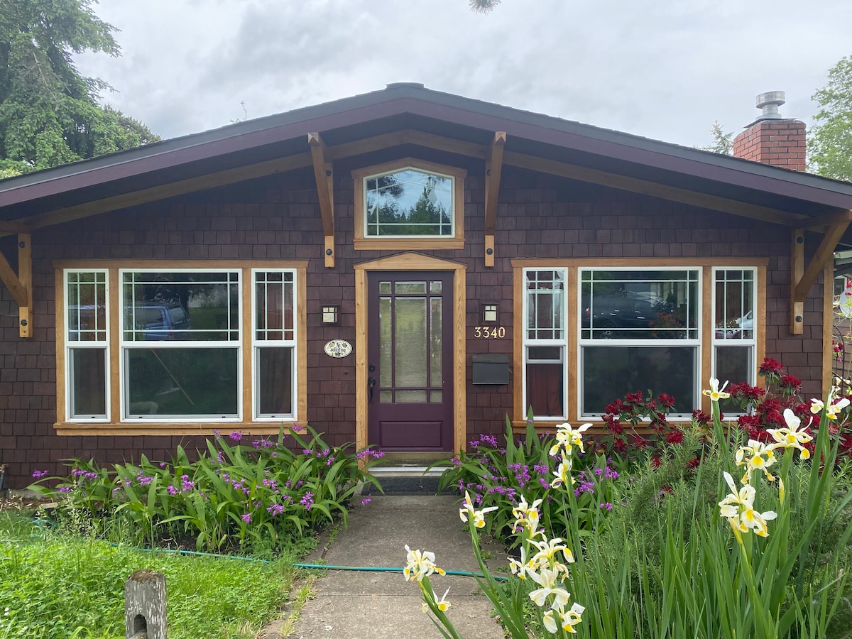 A charming home exterior is displayed, featuring a mix of dark wood shingles and a welcoming front door in a deep hue. Vibrant flowers and lush greenery frame the path leading to the entrance, with large windows allowing natural light to enter.