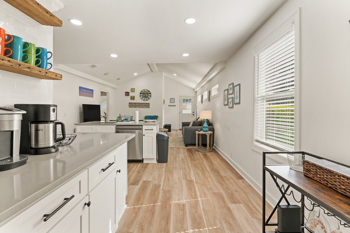 A bright and airy open-plan kitchen and living area is shown, featuring white cabinetry and a stylish countertop. A coffee maker and cups are visible on the kitchen shelves. The living space includes a comfortable seating arrangement with natural light filtering through large windows.