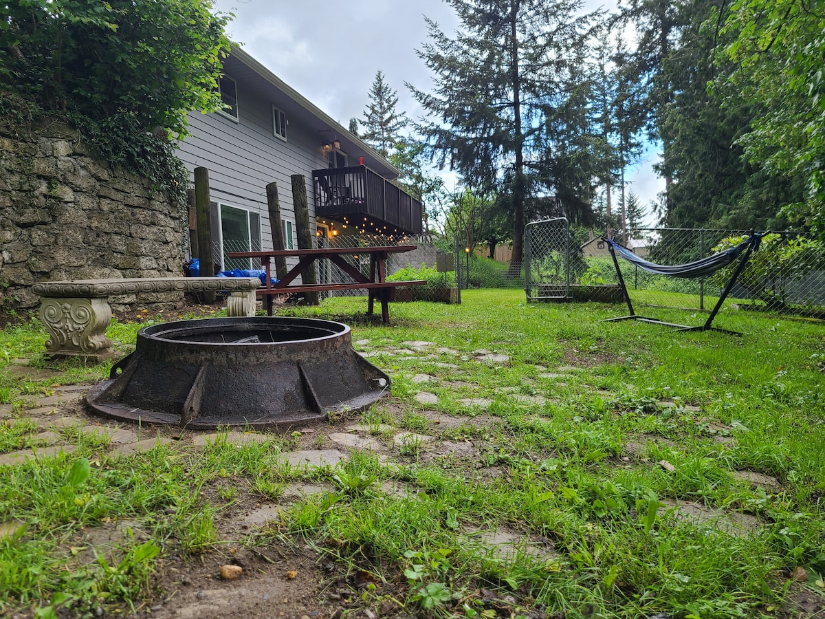 An outdoor space features a fire pit at the forefront, surrounded by stone pavers and lush green grass. A picnic table and a swing hammock are visible, with the exterior of a two-story home and trees providing a serene backdrop.