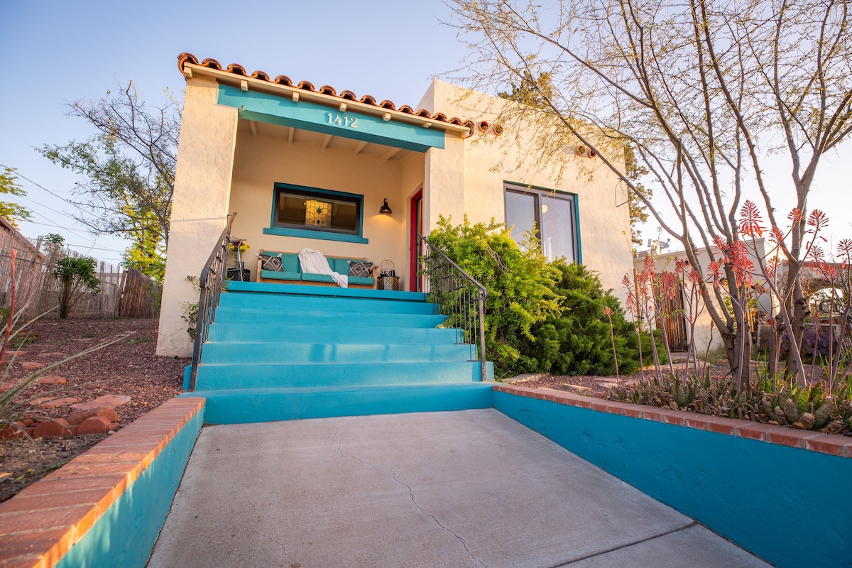 The exterior of a renovated cottage showcases a bright blue front porch with two lounge chairs. Steps lead up to the entrance, framed by various desert plants. The warm hue of the building contrasts with the natural surroundings, creating an inviting entryway.