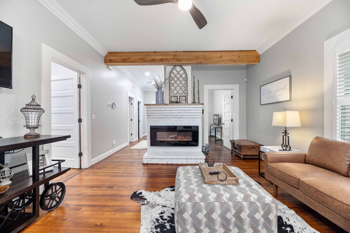 A stylish living area features a modern fireplace set against a whitewashed brick backdrop. A plush sofa is complemented by a decorative pouf and a wooden side table, while natural light enters through nearby windows, enhancing the welcoming atmosphere.
