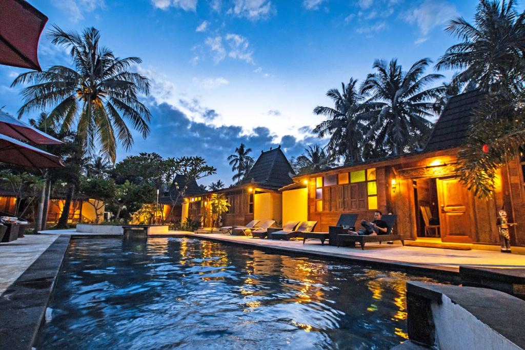A tranquil swimming pool is framed by palm trees and lounge chairs, reflecting the evening sky. The wooden structures provide an inviting backdrop, with warm lighting accentuating their rustic charm. The scene suggests a peaceful retreat, ideal for relaxation.