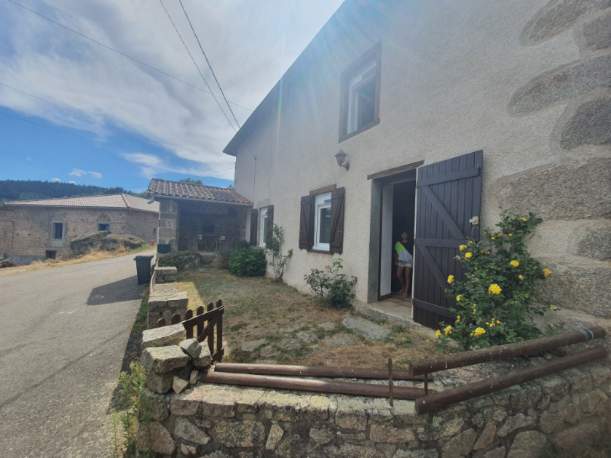 A charming exterior of a house is depicted, featuring a stone facade and wooden accents. A small garden with greenery and yellow flowers is visible. Two windows and a doorway provide access, while a gravel path leads to the street, showcasing a peaceful country setting.