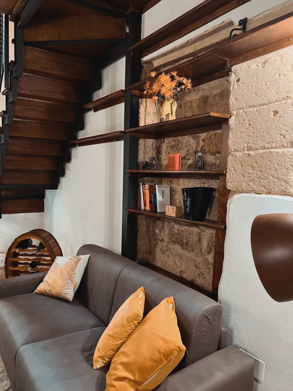 A cozy living area features a chocolate-brown sofa adorned with bright yellow throw pillows. Above the sofa, a rustic wooden shelf holds decorative items and books, while the textured wall behind adds warmth to the space. A staircase with wooden steps is visible in the background.