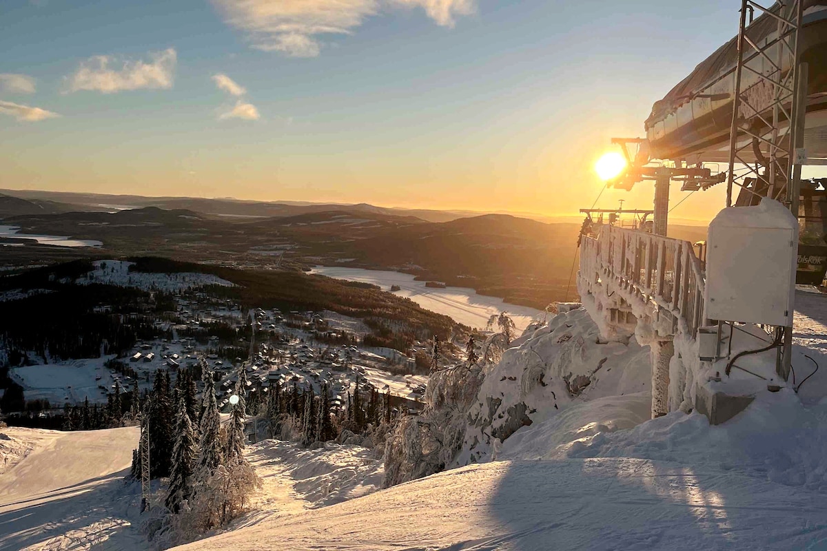 A ski lift stands against a backdrop of a snowy landscape, with the sun setting over the mountainous horizon. Below, a village can be seen nestled among trees and the lake glimmering in the distance, creating a serene winter scene.