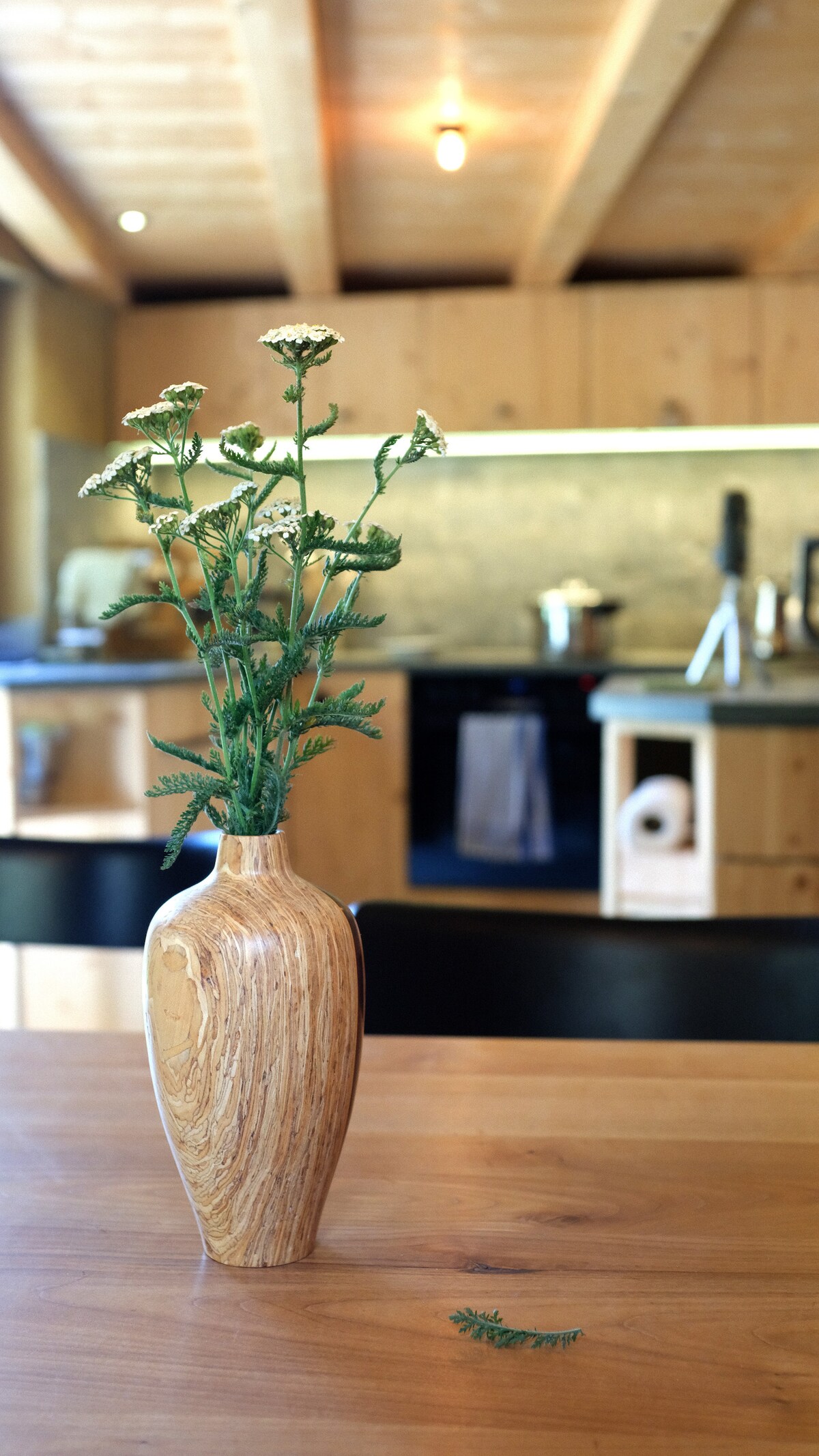 A wooden vase with delicate flowers sits on a polished table. The background reveals a modern kitchen space with wooden cabinetry and subtle lighting that enhances the natural elements of the interior.