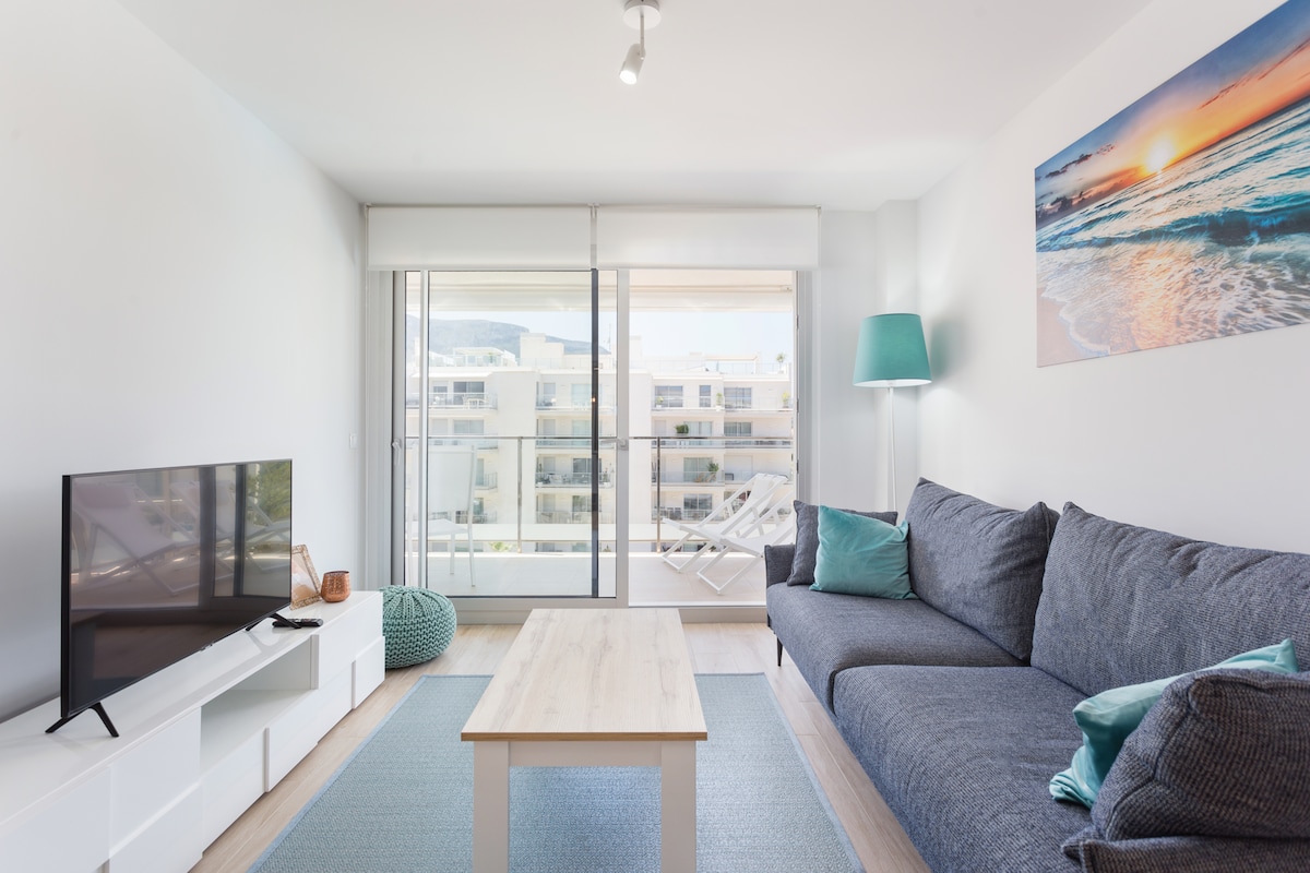 A bright living area features a large gray sofa adorned with teal cushions. A light wooden coffee table is situated in front, while a flat-screen TV rests on a white media unit. Natural light floods the space through glass doors leading to an outdoor terrace.
