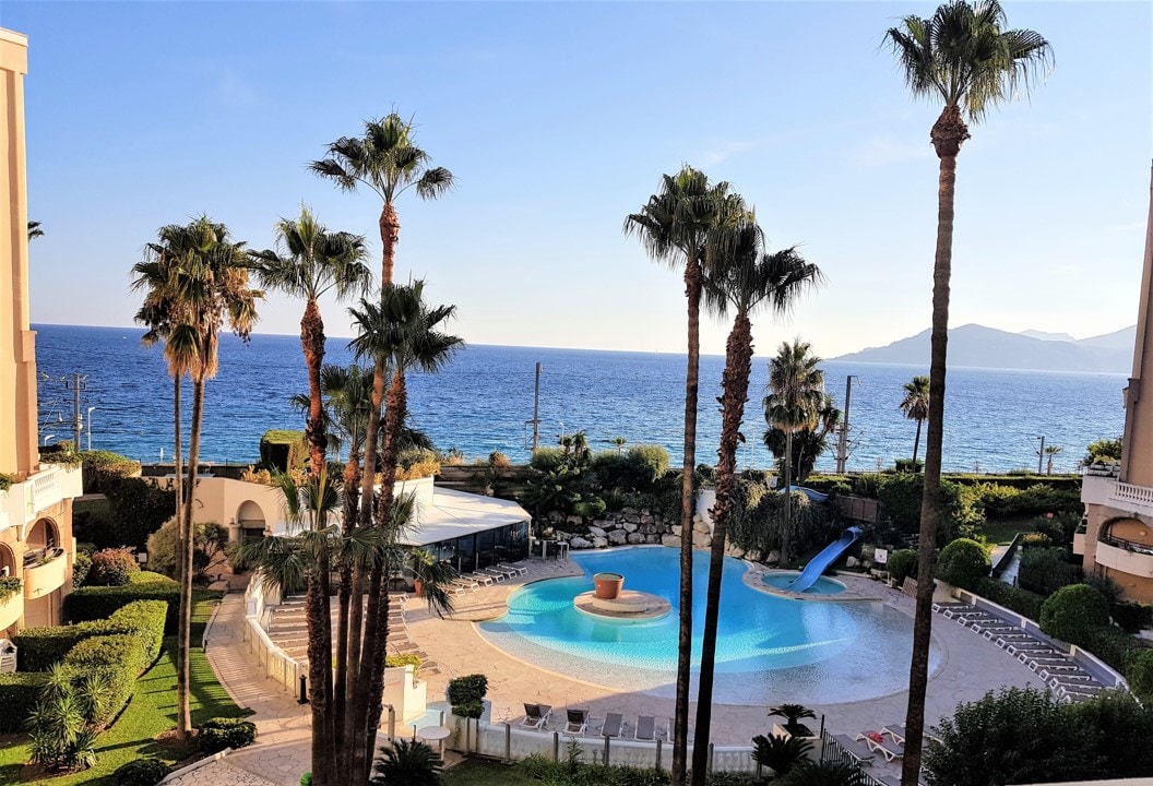 A view of a circular swimming pool surrounded by palm trees, overlooking the sea. The pool features a small fountain and a water slide. Lush greenery and lounge chairs are positioned around the pool area, with the ocean visible in the background under a clear sky.