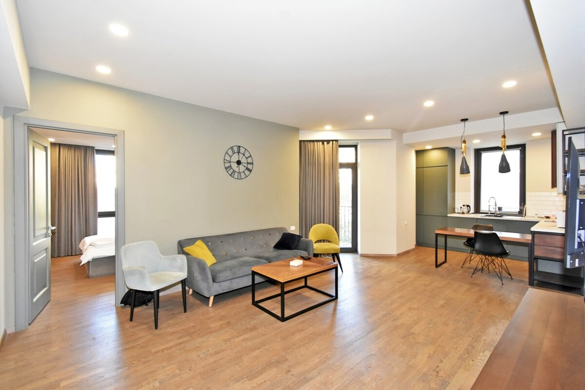 A spacious living area features a gray sofa and an armchair with a yellow accent cushion. A rectangular coffee table is positioned in front of the seating. Natural light fills the room through large windows, accompanied by warm LED lighting. The open kitchen is visible in the background.