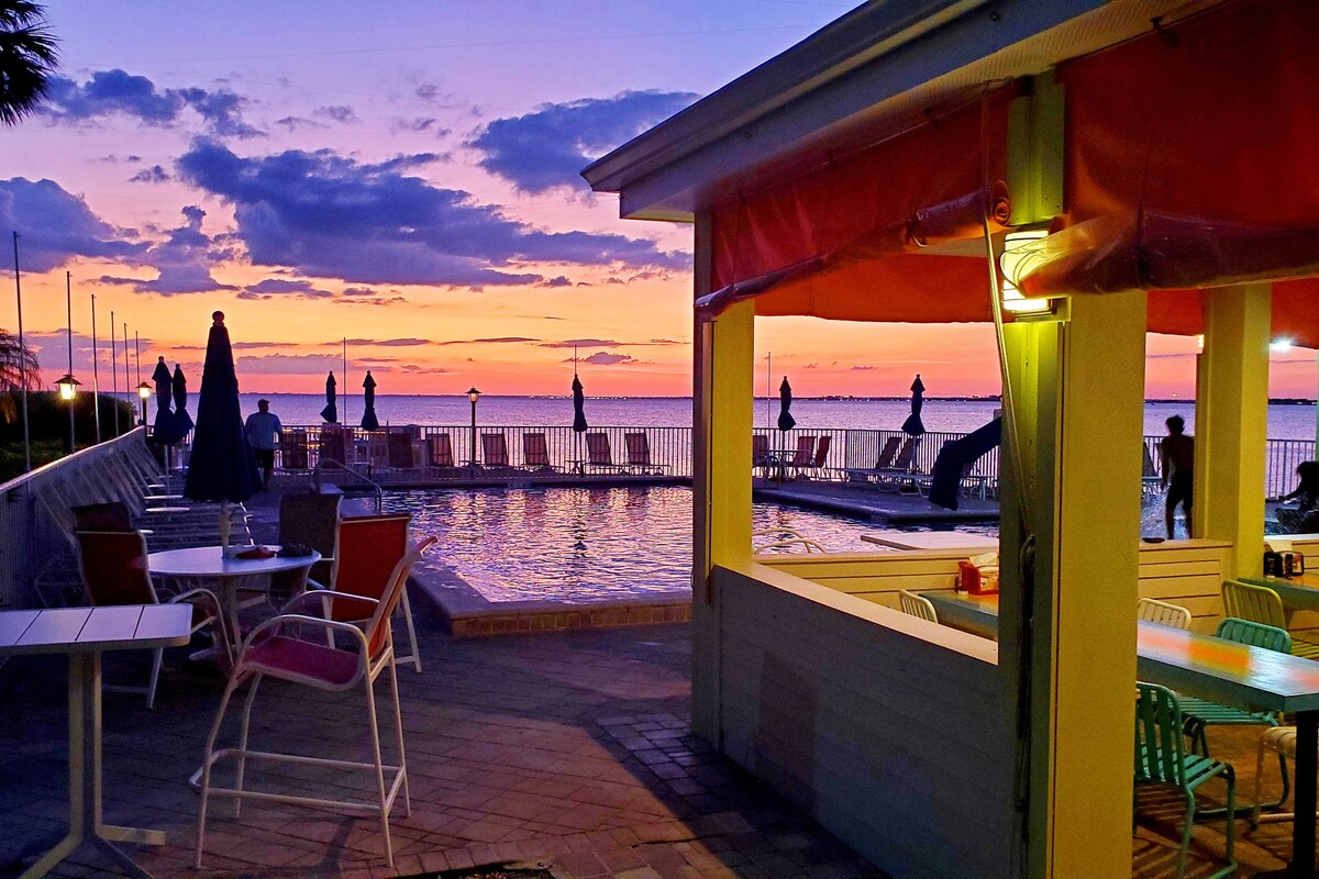 A scenic outdoor area featuring a glowing sunset over the water. In the foreground, there are tables and chairs beside a pool, with large umbrellas providing shade. Soft orange and purple hues fill the sky, reflecting on the water's surface and creating a serene atmosphere.