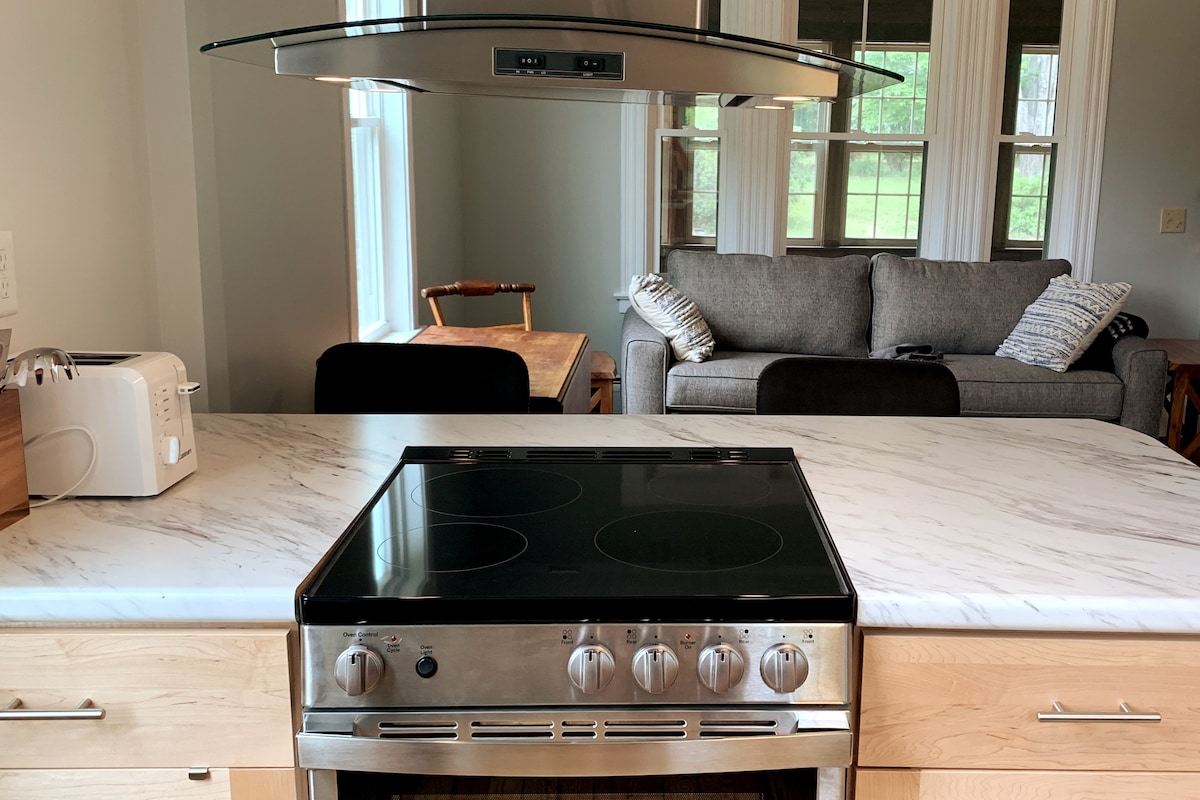 A modern kitchen features a sleek stainless-steel stove integrated into a marble countertop. Behind, a spacious living area is visible with a gray sofa and a wooden dining table. Large windows allow natural light to fill the space.