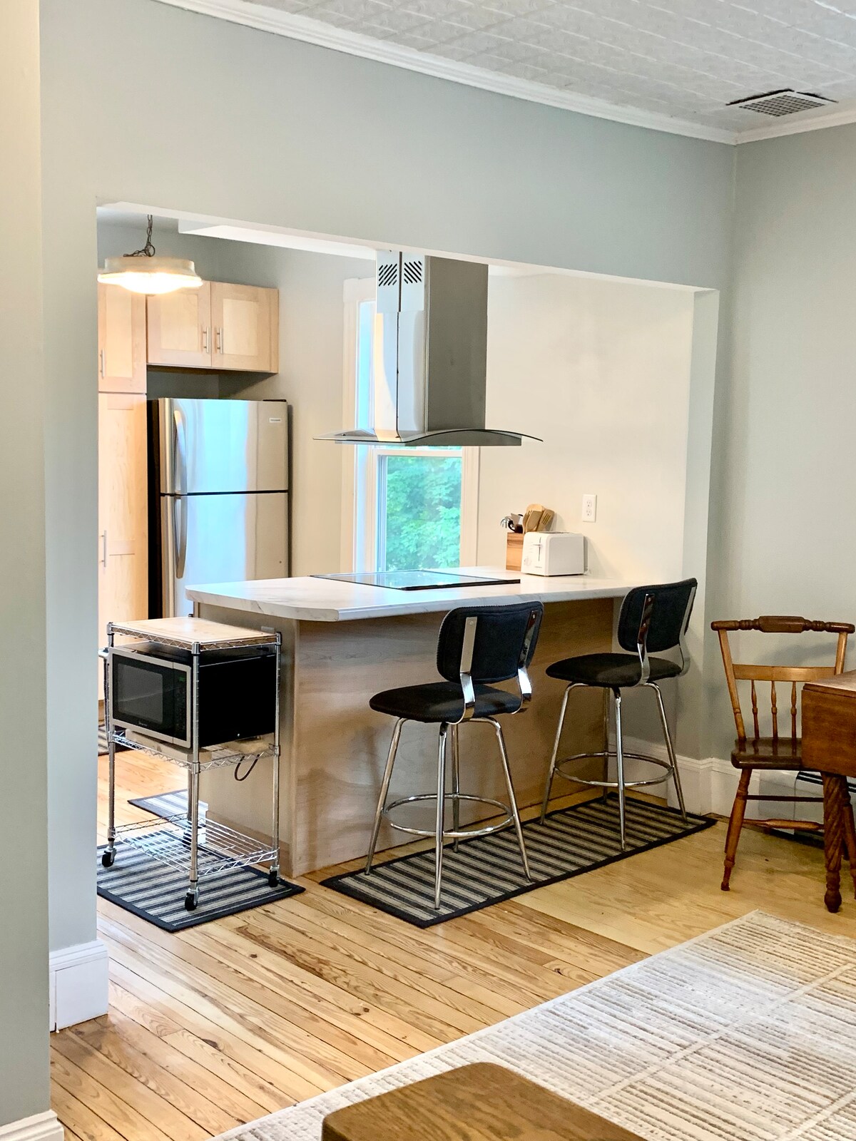 The open kitchen showcases modern stainless-steel appliances, including a refrigerator and hooded range, complemented by a central island with seating. Light wood cabinetry and countertops enhance the space, while large windows provide natural light into the area featuring bamboo mats on the floor.