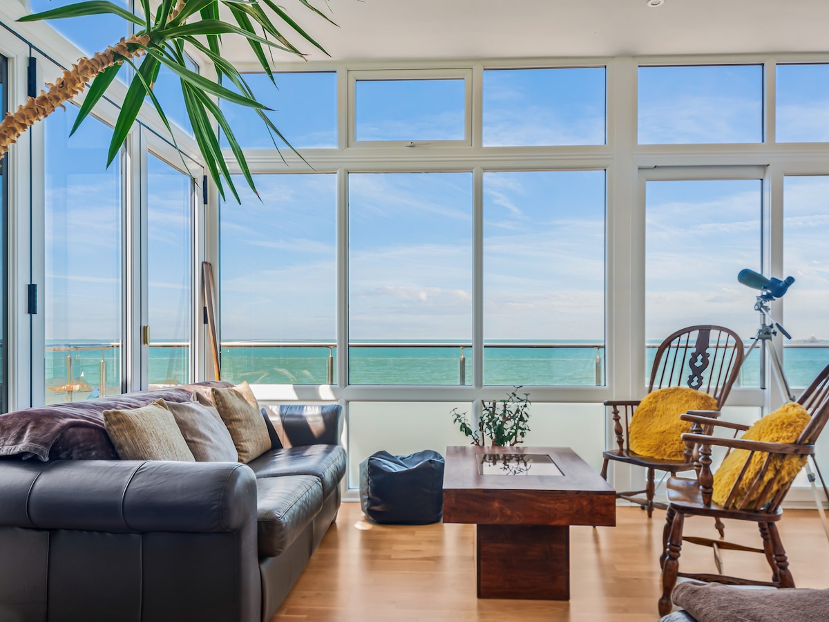 A light-filled living area features large windows framing stunning sea views. Comfortable seating is arranged around a wooden coffee table, complemented by a cozy throw. A telescope is positioned by the glass walls, ideal for enjoying the panoramic scenery.