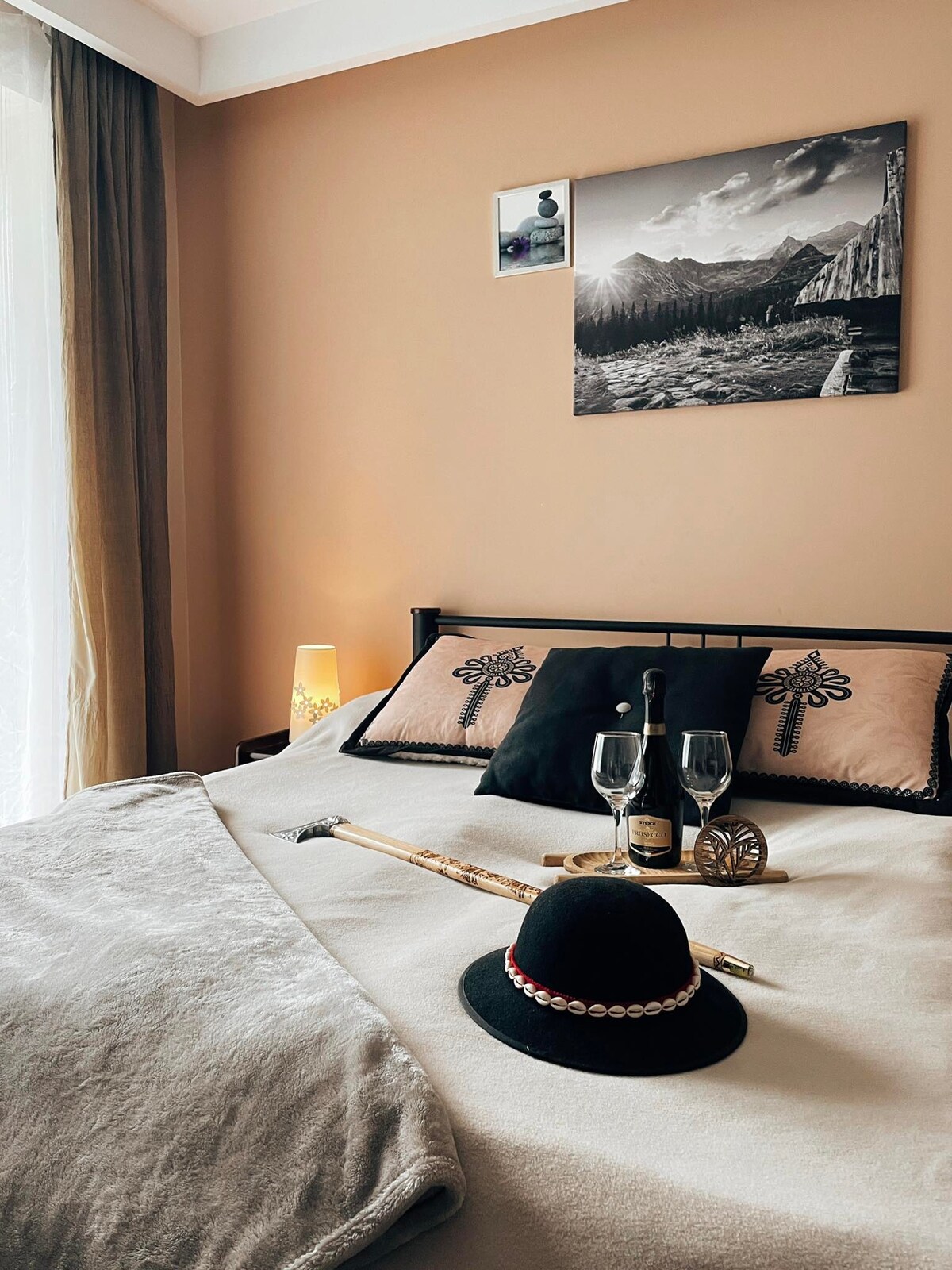 A sleeping area is highlighted with a spacious bed featuring decorative pillows. A soft blanket covers the bed. Two glasses and a bottle are placed on a small table. A hat and a decorative item are also positioned on the bed. Natural light filters through a nearby window.