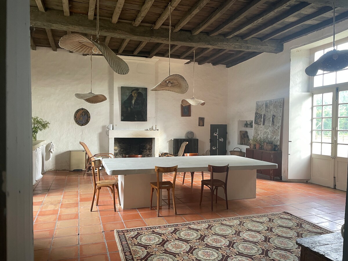 A spacious dining area features a large table surrounded by wooden chairs. Natural light floods in through multiple windows, illuminating the terracotta floor. An art piece and decorative elements are displayed on the white walls, complementing the rustic wooden beams overhead.