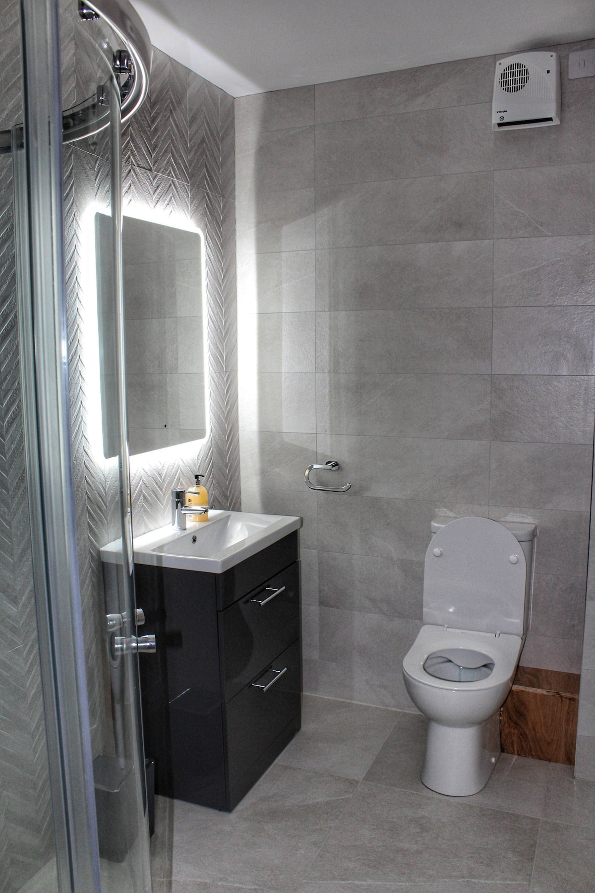 A modern tiled bathroom features a spacious shower enclosed by glass, a sleek vanity with two drawers, and a large illuminated mirror. A white toilet is positioned adjacent to the vanity, with patterned wall tiles adding texture to the design.