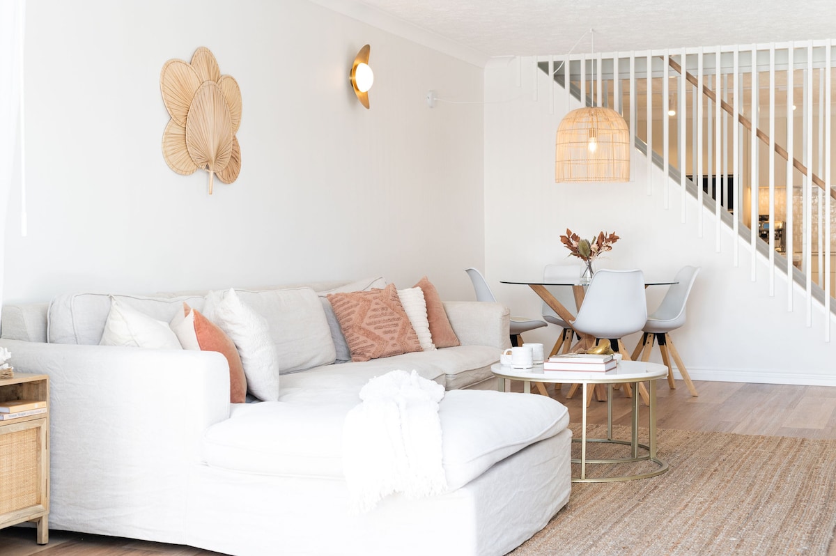 The living area features a sectional sofa adorned with textured cushions and a cozy throw. A round coffee table holds a selection of magazines. A stylish dining table with white chairs is visible in the background, illuminated by a woven pendant light.