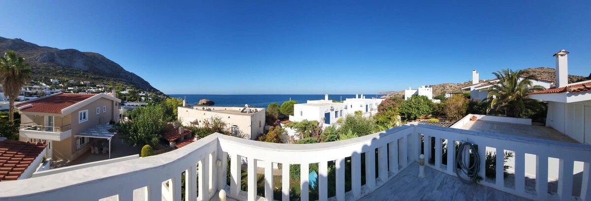 A panoramic view showcases the azure sea beyond well-maintained gardens and whitewashed buildings. Balconies and rooftops are visible in the foreground, surrounded by vibrant greenery under a clear blue sky.