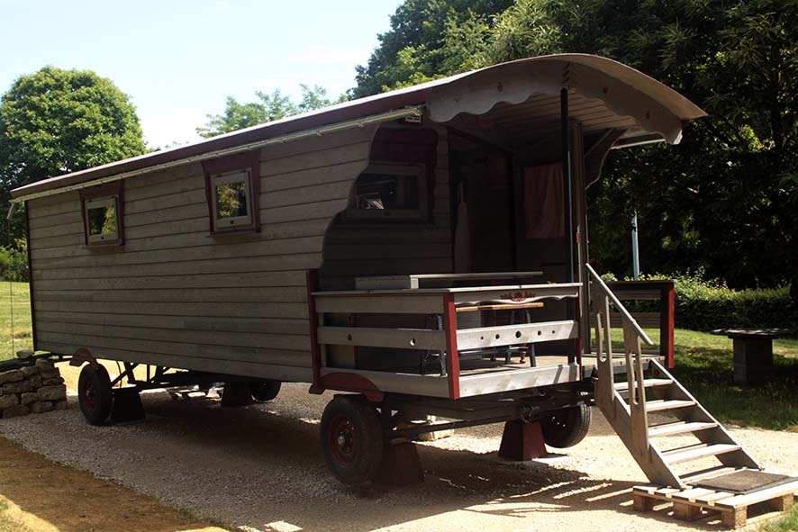 A rustic wooden trailer is set in a natural landscape, featuring a curved roof and large windows. A staircase leads to the entrance, and an outdoor seating area is visible at the front, offering a peaceful escape in a serene environment.