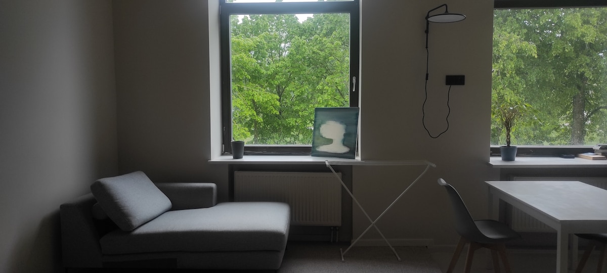 A modern living space features a gray lounge chair positioned beside a large window, allowing for ample natural light. A small table holds an artwork, while greenery is visible through the glass. A minimalistic white table and chair complete the room.
