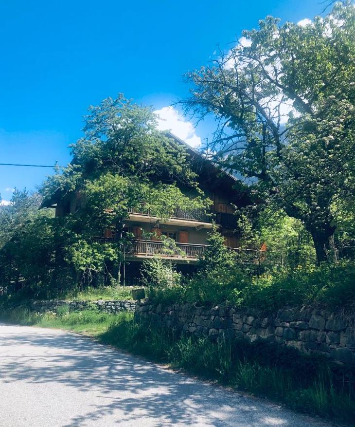 A rustic chalet style building is surrounded by lush greenery and mature trees. The structure features a balcony on the upper floor, providing a place to enjoy the outdoors. A gravel road runs alongside the property, leading to a peaceful mountain setting.