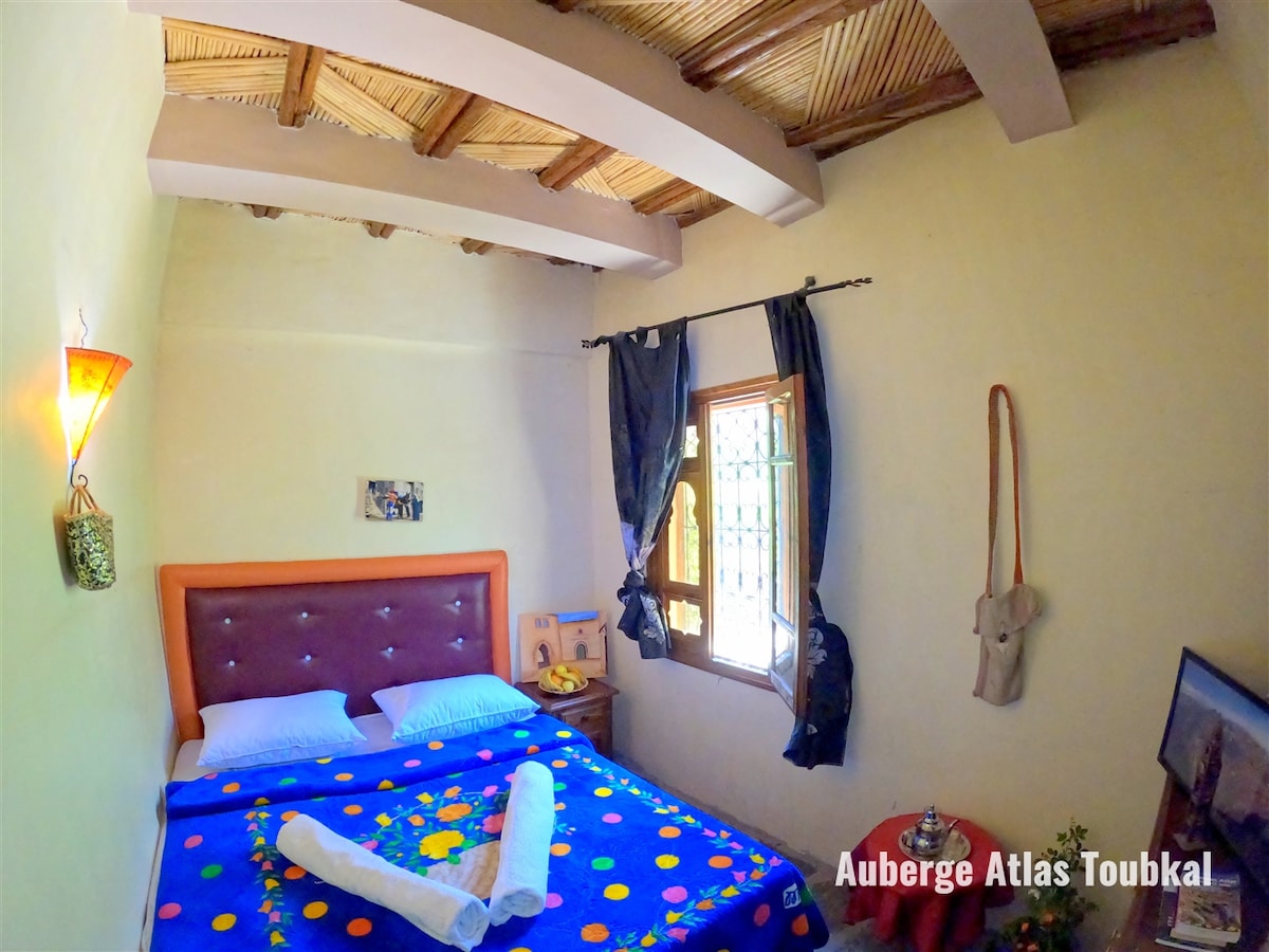 A cozy double room is shown, featuring a colorful floral-patterned bedspread over a spacious bed. Natural light enters through a window with wooden shutters, bordered by dark curtains. The room’s decor includes woven details and wall art, emphasizing a traditional Moroccan style.