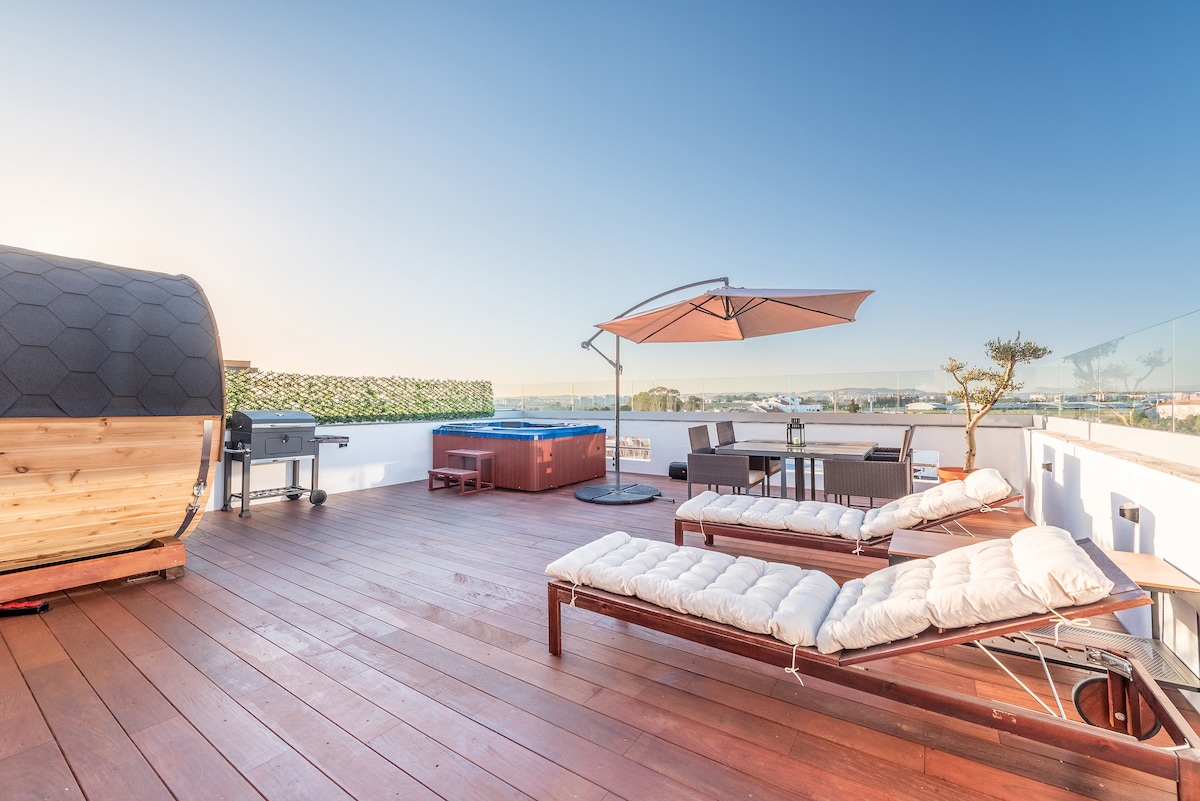 A spacious rooftop terrace features a hot tub, a shaded dining area, and a barbecue grill, accompanied by two sun loungers. The wooden deck provides ample space, while greenery adds a touch of privacy and tranquility to the setting.