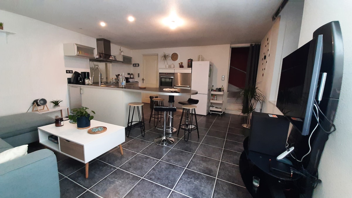 An open living area features a modern kitchen with stainless steel appliances and a spacious island. Seating includes a gray sofa, black stools at the island, and a low coffee table with plants. A flat-screen TV is positioned in the corner, with a staircase visible in the background.