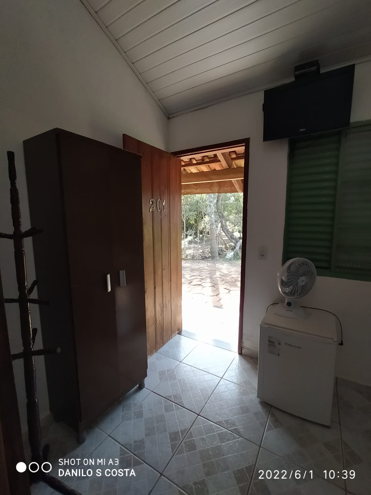 A simple entrance area is highlighted, featuring a wooden door opening to an outdoor space. A dark wardrobe and a small refrigerator are positioned against the wall. A ceiling fan is visible, and light fills the space through a nearby window with green shutters.