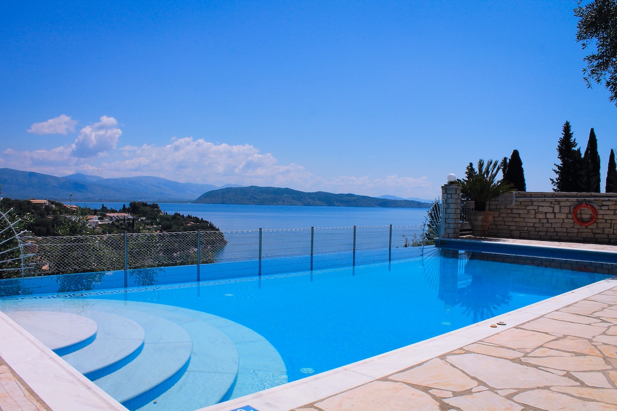 A serene infinity pool is visible, featuring gentle steps leading into the clear blue water. The surrounding area includes a stone patio and panoramic views of the coastline and hills, framed by lush greenery and a clear sky.